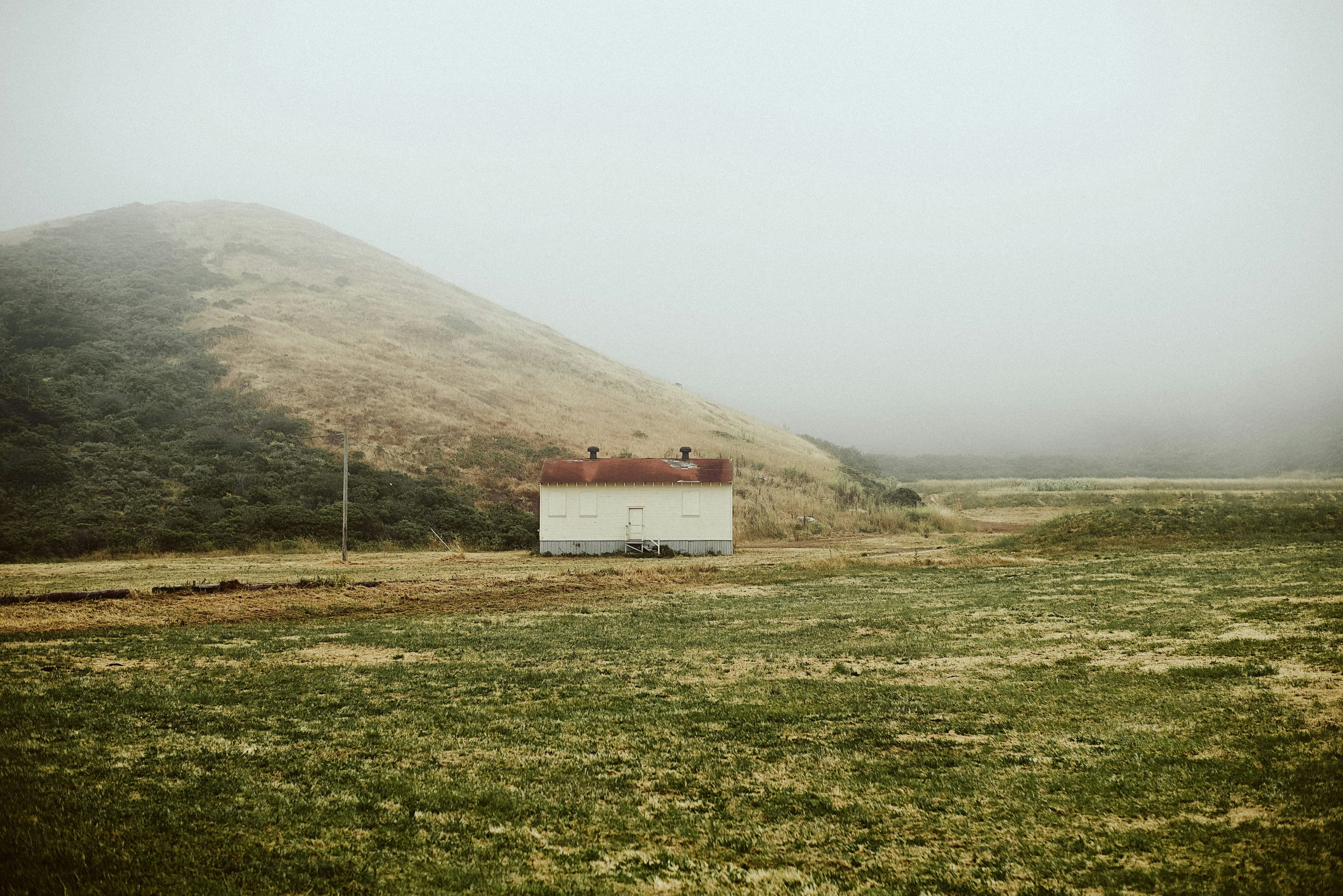 A small white building in a misty green field beneath a hill